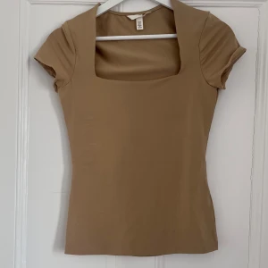 Beige topp från H&M - Säljer en stilren beige topp från H&M med korta ärmar och fyrkantig halsringning. Perfekt för en enkel och elegant look. Toppen har en tight passform som framhäver figuren.