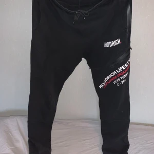 Svarta mjukisbyxor från Hoodrich - Snygga svarta mjukisbyxor från Hoodrich med tryck i vitt och rött. Byxorna har en bekväm passform med snörning i midjan och en dragkedja på sidan. Perfekta för en avslappnad stil.