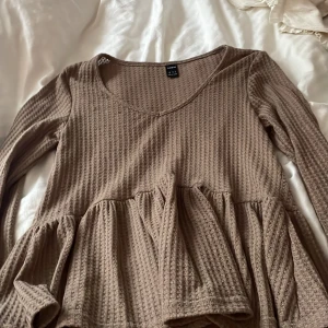 Brun långärmad topp från Shein - Säljer en brun långärmad topp från Shein med en vacker volangdetalj i midjan. Toppen har en ribbad textur och en rundad halsringning. Perfekt för en stilren och bekväm look.