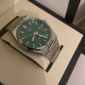 Grön klocka från Tissot - Säljer en stilren Tissot-klocka med grön urtavla och silverfärgad metallarmband. Klockan har datumvisning och kommer i originalförpackning med certifikat. Perfekt för den som vill ha en elegant accessoar.