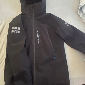 Svart jacka från Sail Racing - Säljer en svart jacka från Sail Racing med dragkedja och huva. Jackan har en stilren design med logotypdetaljer på ärmen och bröstet. Perfekt för kyligare dagar.