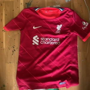 Röd Liverpool fotbollströja - Säljer en röd Liverpool fotbollströja från Nike med korta ärmar. Tröjan har vita detaljer och klubbens emblem på bröstet. På baksidan finns namnet och numret 'M. Salah 11'. Perfekt för fans av laget!