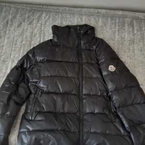 Svart dunjacka från Moncler - Säljer en snygg svart dunjacka från Moncler med hög krage och dragkedja framtill. Jackan har två praktiska fickor med dragkedjor och Monclers logga på ärmen. Perfekt för kyliga dagar!