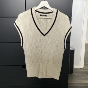 Vit stickad väst från Gina Tricot - Säljer en stilren vit stickad väst från Gina Tricot med svarta detaljer. Västen har en v-ringad design och ett klassiskt flätmönster. Den är stor i storleken men är gjord för att vara lite baggy. Perfekt för lager-på-lager-stil. Väldigt skön och mjuk, håller även värmen bra! .