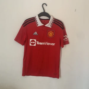  Manchester United fotbollströja 2022  - Säljer en röd Manchester United fotbollströja från Adidas med vit krage och svarta detaljer. Tröjan har korta ärmar och klubbens emblem på bröstet. Perfekt för fans som vill visa sitt stöd på matchdagar! ⚽