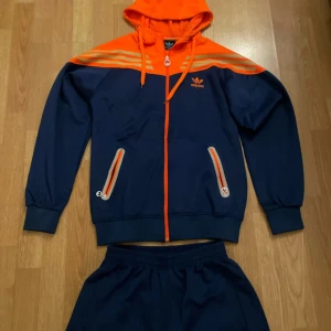 Blå och orange hoodie från Adidas - Säljer en snygg blå hoodie från Adidas med orangea detaljer och dragkedja. Tröjan har en klassisk Adidas-logga på bröstet och huva med dragsko. Byxorna kommer med också.