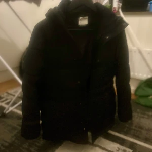 Svart pojke pufferjacka med huva - Säljer en svart pufferjacka med huva. Jackan har långa ärmar och en dragkedja framtill. Perfekt för kyligare dagar.