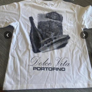 Vit t-shirt från Yacht Club - Säljer en stilren vit t-shirt från Yacht Club med tryck på både fram- och baksida. Framsidan har en diskret logga medan baksidan pryds av ett större motiv med texten 'Dolce Vita Portofino'. Perfekt för en avslappnad och elegant look.