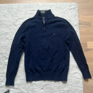 Mörkblå tröja med dragkedja från Tommy Hilfiger - Säljer en stilren mörkblå tröja från Tommy Hilfiger med dragkedja vid halsen. Tröjan har långa ärmar och en liten logotyp på bröstet. Perfekt för en avslappnad och snygg look.