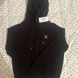 Svart hoodie från Calvin Klein - Säljer en stilren svart hoodie från Calvin Klein med en liten CK-logga på bröstet. Den har en klassisk huva med dragsko och långa ärmar. Perfekt för en avslappnad look.