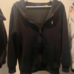 Svart hoodie från Polo Ralph Lauren - Säljer en stilren svart hoodie från Polo Ralph Lauren med dragkedja och det klassiska broderade logotypen på bröstet. Perfekt för en avslappnad look. Den har långa ärmar och en praktisk huva.
