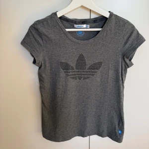 Adidas grå t-shirt, storlek 36 - Grå t-shirt från Adidas med en stor Adidas-logga gjord av små svarta stenar på framsidan.  T-shirten har korta ärmar och en rund halsringning. En liten Adidas-logga finns även på vänster nederkant. Mycket bra skick. Tvättad. Djurfritt och rökfritt hem.