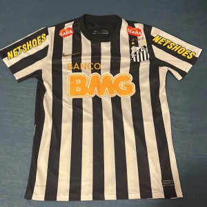 Randig fotbollströja från Nike - Säljer en randig fotbollströja från Nike fc santos med svart och vit design. Tröjan har korta ärmar och flera sponsormärken, inklusive BMG och Netshoes. Perfekt för sommaren om man vill sticka utt den är otroligt sällsynt och äkta men den är en del gammal men sparsamt använd! 