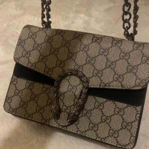 Mönstrad axelväska från G u c c i - Säljer en stilren axelväska med det klassiska GG-mönstret. Väskan har en elegant kedjerem och ett dekorativt spänne framtill. Perfekt för att ge din outfit en lyxig touch.