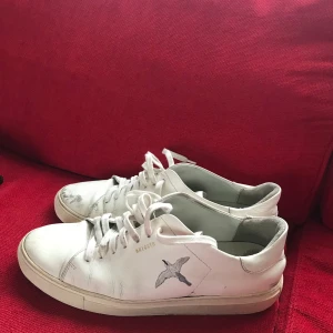 Vita sneakers från Axel Arigato - Snygga vita sneakers från Axel Arigato med en stilren design och fågelmotiv på sidan. Skorna har snörning och en rund tå, vilket ger en klassisk look. Perfekta för en avslappnad stil.