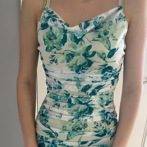 Blommig klänning med gröna nyanser - Säljer en vacker blommig klänning med gröna nyanser på en vit bakgrund. Klänningen har en tight passform och är ärmlös. Perfekt för sommarens festligheter!