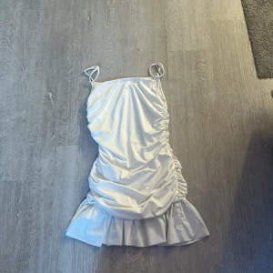 Vit klänning med volang - Säljer en stilren vit klänning med smala axelband och volangdetalj vid fållen. Klänningen har en åtsittande passform med rynkade detaljer på sidorna som ger en snygg effekt. Perfekt för sommarens fester!