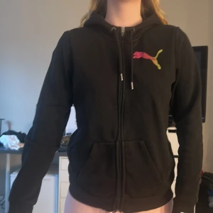 Svart hoodie från Puma - Säljer en svart hoodie från Puma med dragkedja och färgglatt logotyptryck på bröstet. Tröjan har långa ärmar och en bekväm passform. Perfekt för en avslappnad stil.