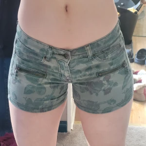 Lågmidjade gröna shorts - Snygga gröna shorts från HM med blommigt mönster och coola dragkedjor framtill. Köpt för säkert 10+ år sedan. De har en låg midja och är tillverkade i ett mjukt, stretchigt material.