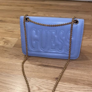 Blå axelväska från Guess - Snygg blå axelväska från Guess med kedjeaxelrem i guld. Väskan har en stilren design med Guess-logga på framsidan och ett stort G på baksidan. Perfekt för att ge din outfit en färgklick!