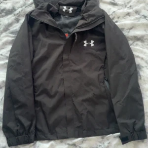 Svart vindjacka från Under Armour - Säljer en svart vindjacka från Under Armour med huva och dragkedja. Jackan har långa ärmar och elastiska muddar. Perfekt för utomhusaktiviteter med sin lätta och funktionella design.