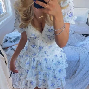 Blommig volangklänning med puffärm - Superfin vit kortklänning med blått blommönster på. Klänningen har v-ringning, volanger och puffärmar. Säljer då den inte kommit till användning🩵