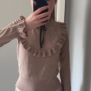 Beige stickad tröja med volang och knytband - Supersöt beige stickad tröja med volangdetaljer runt bröstet och halsen samt ett svart knytband framtill. Tröjan har långa ärmar och ribbade muddar. Perfekt för dig som gillar romantiska detaljer och mjuka material.