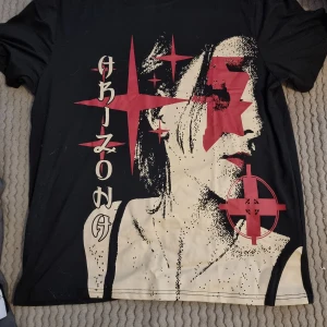 Svart t-shirt med grafiskt tryck - Cool svart t-shirt med stort grafiskt tryck av ett ansikte i beige och rött, samt röda stjärnor. T-shirten har rund hals och korta ärmar, perfekt för en edgy streetstyle-look. 