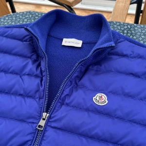 Blå dunväst från Moncler - Snygg blå dunväst från Moncler med quiltad design och dragkedja framtill. Västen har ribbade detaljer vid krage och nederkant samt två praktiska fickor med dragkedja. Perfekt för lager-på-lager och stilren look.