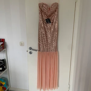 Glittrig axelbandslös långklänning i ljusrosa - Säljer en superfin axelbandslös långklänning i ljusrosa med glittrig paljettöverdel och flowig kjol i chiffongliknande material. Perfekt för bal eller fest! Klänningen har en snygg sweetheart-ringning och markerad midja. Prislappen är på!