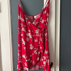 Röd blommig omlottklänning med volang - Säljer en röd omlottklänning med blommigt mönster och volangdetaljer. Klänningen har smala axelband och knyt i midjan för en fin passform. Perfekt för sommarens alla tillfällen!