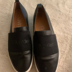 Svarta slip-on skor från Calvin Klein - Säljer ett par svarta slip-on skor från Calvin Klein med bred resår över foten och rund tå. Skorna har ovandel i skinn och en vit platt sula. Perfekta för en stilren och enkel look.
