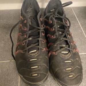 Nike Air Vapormax Plus svarta sneakers - Nike Air Vapormax Plus sneakers i svart med röda detaljer och synlig luftdämpning i sulan. Skorna har snörning, rund tå och en unik, futuristisk design med syntetmaterial och tydlig Nike-logga på sidan.