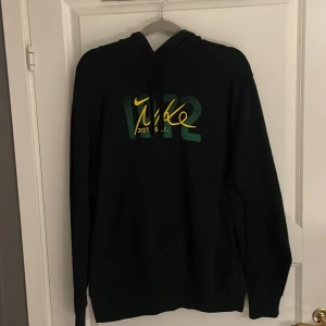 Svart hoodie från Nike med grafiskt tryck - Svart hoodie från Nike med grönt och gult grafiskt tryck framtill och stort tryck på ryggen. Huva och långa ärmar. Mjuk insida och klassisk passform. Perfekt för dig som gillar sportig stil. Skick 10/10 då den aldrig är andvänd! Bara att höra av sig vid frågor 