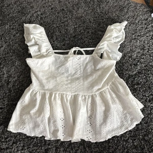 Vit broderad volangtopp från SHEIN - Superfin vit topp från SHEIN med broderade detaljer och volang på axelbanden. Toppen har knytning i ryggen och är tillverkad i polyester med elastan. Perfekt för sommardagar och ger en romantisk look.