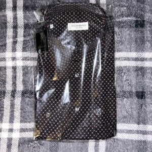 Mönstrad skjorta från Yves Saint Laurent - Säljer en exklusiv skjorta från Yves Saint Laurent pour homme. Skjortan är svart med ett småmönstrat tryck i beige och har klassisk krage samt knäppning framtill. Perfekt för dig som vill sticka ut med stil.
