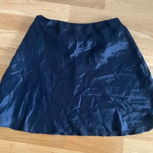 Svart satin kjol från Chiquelle - Säljer en svart, glansig kjol från Chiquelle i satinmaterial. Kjolen har en enkel A-linjeform och är kort i modellen. Perfekt för dig som gillar stilrena plagg med lite extra glans.