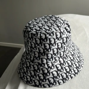 Vit och svart bucket hat med monogrammönster - Snygg bucket hat i vitt och svart med ett tätt monogrammönster över hela hatten. Hatten har klassisk form med nedvikt brätte och är tillverkad i ett mjukt tygmaterial. Perfekt accessoar för att lyfta din stil.