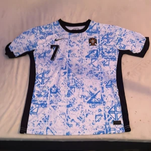 Portugal fotbollströja Nike Dri-Fit - Säljer en Portugal fotbollströja från Nike i Dri-Fit material. Tröjan är vit med blått grafiskt mönster, svarta detaljer på sidorna och runt kragen. Nummer 7 på bröstet och Portugals landslagsmärke. Perfekt för fotbollsfans!