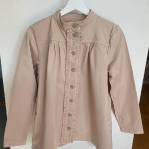 Beige vintagejacka  med volangkant från Rodebjer stl S - Säljer en beige vintagejacka från Rodebjer i storlek S. Jackan har långa ärmar, knappar framtill och en fin volangkant längs knappraden. Perfekt för dig som gillar stilrena detaljer och mjuka material. Jackan är i bomull/linnemix. 