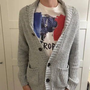 Ljusgrå stickad kofta från Divided - Säljer en ljusgrå stickad kofta från Divided med grovstickat mönster, stora knappar framtill och två fickor. Koftan har ribbade muddar och förstärkta armbågar i ljusare tyg. Perfekt för lager-på-lager-stil.