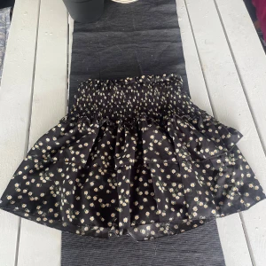 Svart blommig kjol från SHEIN - Supersöt svart kortkjol från SHEIN med vita och gula prästkragar. Kjolen har smock i midjan och volangig nederdel som ger en luftig känsla. Perfekt för dig som gillar blommiga mönster och vill ha något bekvämt och trendigt.