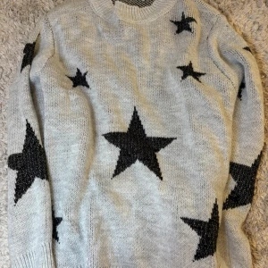 Ljusgrå stickad tröja med svarta stjärnor - Säljer en ljusgrå stickad tröja med stora svarta stjärnor. Tröjan har rund halsringning och långa ärmar. Perfekt för dig som gillar en cool och mysig stil.
