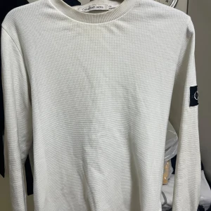 Vit långärmad tröja från Calvin Klein Jeans - Säljer en stilren vit långärmad tröja från Calvin Klein Jeans i storlek XS. Tröjan har ett diskret strukturmönster och en svart logotyp-patch på ena ärmen. Perfekt för en clean och enkel look.