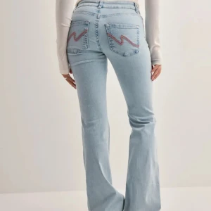 Ljusblå bootcut jeans med broderade bakfickor - Snygga ljusblå jeans med bootcut passform och slitningar på båda knäna. Bakfickorna har ett broderat zigzag-mönster i rosa. Jeansen har klassisk femficksdesign och stängs med dragkedja och knapp.