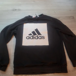 Svart sweatshirt från Adidas - Svart sweatshirt från Adidas med stor vit ruta och klassisk Adidas-logga på bröstet. Tröjan har rund hals och långa ärmar. Perfekt för en sportig och avslappnad stil. Tvättas innan fraktning 