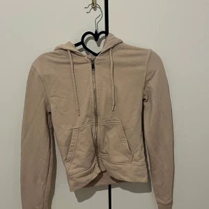 Beige hoodie med dragkedja från H&M - Säljer en beige hoodie från H&M Basic i storlek XS. Tröjan har dragkedja framtill, huva med snören och två fickor. Perfekt för en avslappnad stil och enkel att matcha med andra plagg.