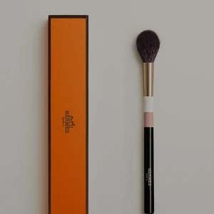 Sminkborste från Hermès - Lyxig sminkborste från Hermès med svart och vit skaft, guldfärgad metall och mjuka, mörka borst. Perfekt för applicering av puder, rouge eller highlighter. Kommer i exklusiv orange Hermès-förpackning.