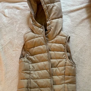 Beige dunväst med huva från ONLY - Säljer en beige dunväst från ONLY med huva och dragkedja framtill. Västen är quiltad och har en enkel, stilren design som passar perfekt till lager-på-lager-look. Perfekt för kyliga dagar när du vill hålla dig varm men ändå röra dig fritt.