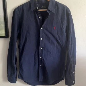 Mörkblå skjorta från Polo Ralph Lauren - Säljer en klassisk mörkblå skjorta från Polo Ralph Lauren i slim fit. Skjortan har vita knappar och den ikoniska röda broderade loggan på bröstet. Perfekt för en stilren och tidlös look.